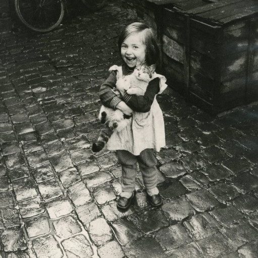Photographie en noir et blanc d'une petite fille debout sur des pavés mouillés, tenant un chaton dans les bras. Elle rit à pleines dents, les yeux plissés, vêtue d'un tablier et de chaussures à boucle. À l'arrière-plan, une caisse en bois et une roue de vélo.